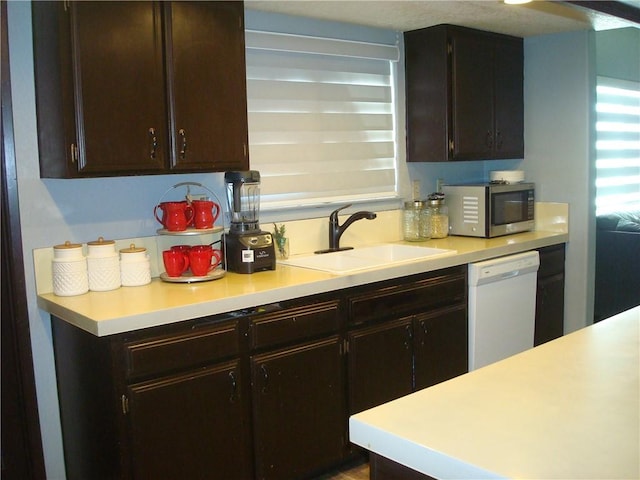 kitchen with dark brown cabinetry, stainless steel microwave, white dishwasher, light countertops, and a sink