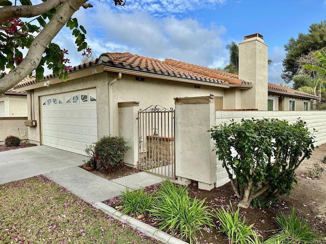 view of side of property with an attached garage, a tile roof, concrete driveway, stucco siding, and a chimney