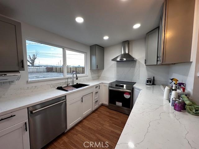 kitchen with stainless steel appliances, a sink, wall chimney range hood, light stone countertops, and dark wood-style floors