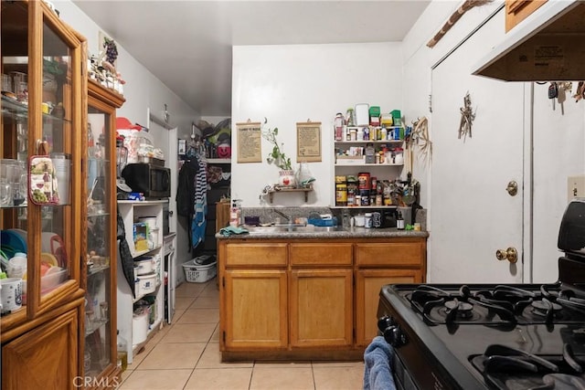 kitchen featuring light tile patterned floors, under cabinet range hood, brown cabinets, dark countertops, and range with gas cooktop