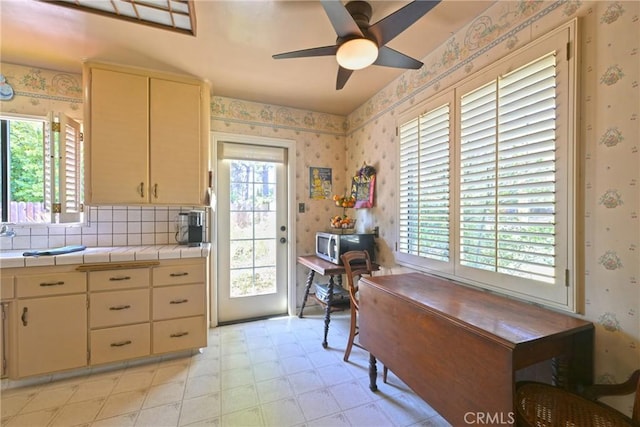 kitchen with tile counters, stainless steel microwave, cream cabinets, ceiling fan, and wallpapered walls