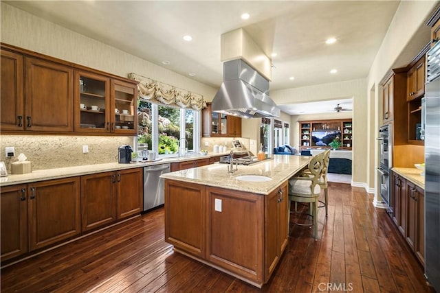 kitchen featuring stainless steel appliances, dark wood-type flooring, glass insert cabinets, a kitchen island with sink, and light stone countertops