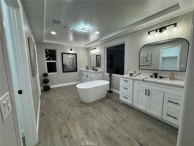 full bathroom with a tray ceiling, a soaking tub, two vanities, wood finished floors, and baseboards