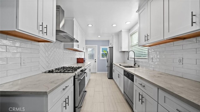 kitchen featuring light stone counters, a sink, white cabinetry, appliances with stainless steel finishes, and wall chimney exhaust hood