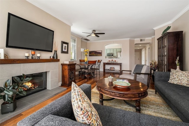 living area with wood finished floors, visible vents, a ceiling fan, ornamental molding, and a tiled fireplace