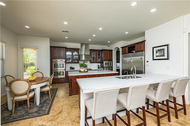 kitchen featuring built in appliances, a kitchen island with sink, a peninsula, a sink, and wall chimney range hood