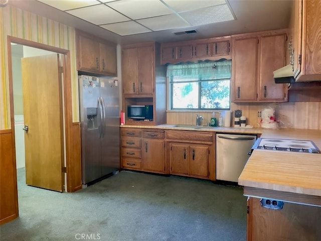 kitchen with brown cabinets, stainless steel appliances, a sink, under cabinet range hood, and wallpapered walls