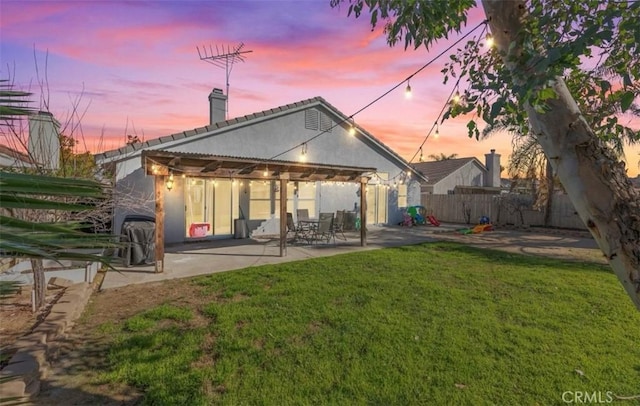 back of property at dusk featuring a yard, a chimney, stucco siding, a patio area, and fence