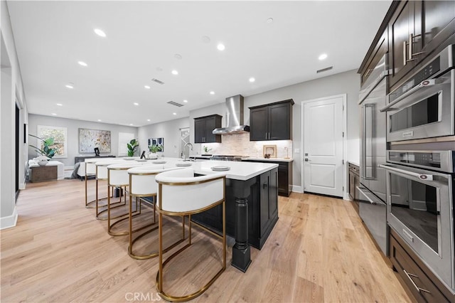 kitchen featuring light wood finished floors, a kitchen breakfast bar, light countertops, stainless steel double oven, and wall chimney range hood