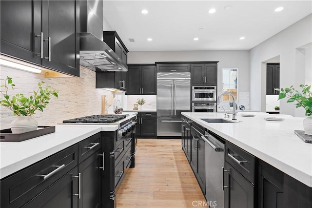 kitchen with wall chimney exhaust hood, stainless steel appliances, dark cabinetry, light countertops, and a sink