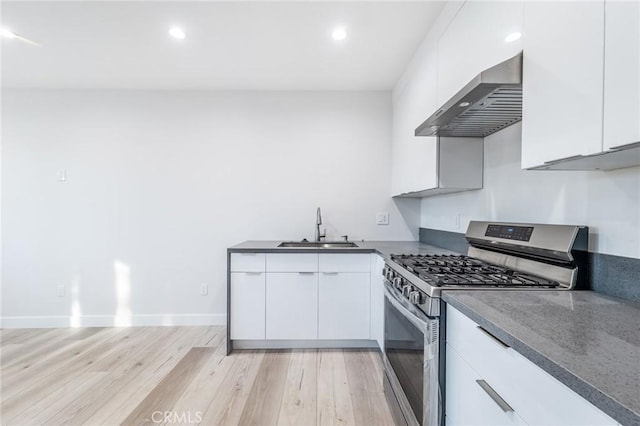 kitchen with a sink, stainless steel gas range oven, white cabinetry, wall chimney range hood, and light wood-type flooring