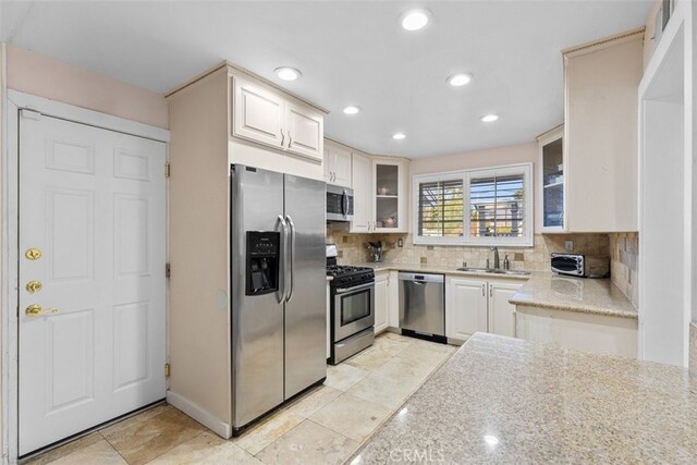 kitchen with tasteful backsplash, glass insert cabinets, light stone countertops, stainless steel appliances, and a sink