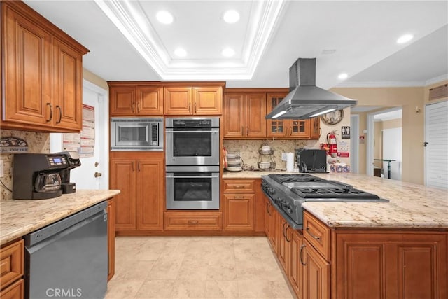 kitchen with island range hood, brown cabinetry, a raised ceiling, appliances with stainless steel finishes, and a peninsula