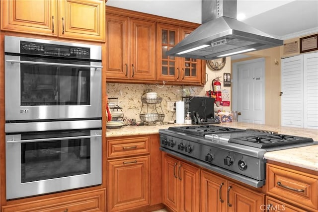 kitchen with stainless steel appliances, brown cabinets, light stone countertops, island exhaust hood, and glass insert cabinets