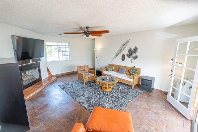 living room with a fireplace with raised hearth, ceiling fan, a textured ceiling, and baseboards