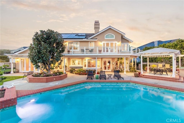 back of property at dusk with french doors, roof mounted solar panels, a patio area, a balcony, and a pergola