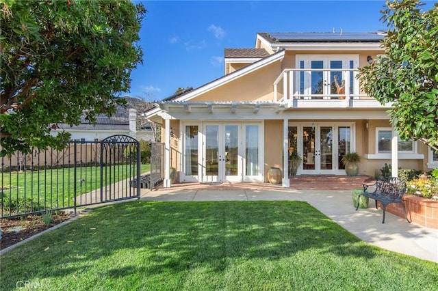 rear view of house with french doors, stucco siding, a lawn, roof mounted solar panels, and fence