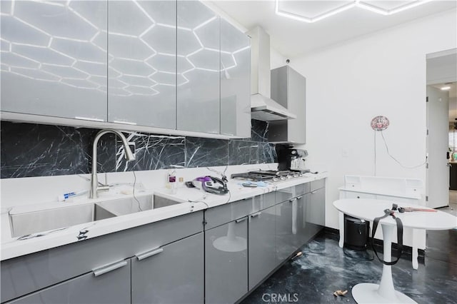 kitchen featuring tasteful backsplash, wall chimney exhaust hood, gray cabinetry, stainless steel gas cooktop, and a sink