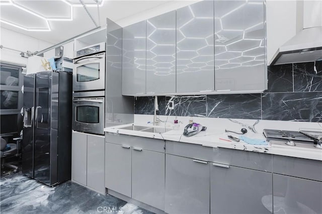 kitchen featuring wall chimney range hood, stainless steel double oven, backsplash, and gray cabinetry