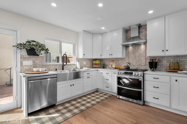 kitchen with stainless steel appliances, light countertops, white cabinets, a sink, and wall chimney exhaust hood