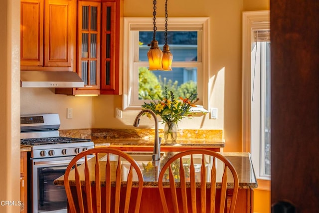 kitchen featuring hanging light fixtures, brown cabinetry, glass insert cabinets, under cabinet range hood, and stainless steel gas range oven