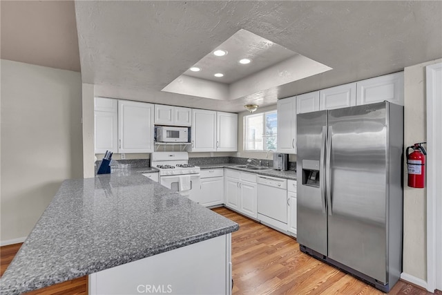 kitchen with a raised ceiling, a peninsula, white appliances, white cabinets, and light wood-style floors