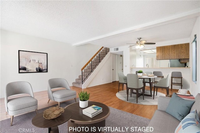 living area with stairway, a ceiling fan, a textured ceiling, wood finished floors, and beamed ceiling