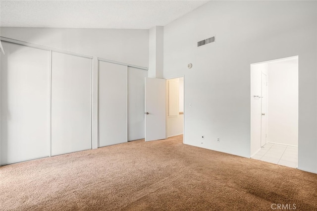 unfurnished bedroom with two closets, visible vents, light carpet, a textured ceiling, and high vaulted ceiling