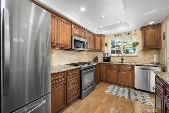 kitchen featuring a tray ceiling, stainless steel appliances, brown cabinetry, light wood-style floors, and a sink