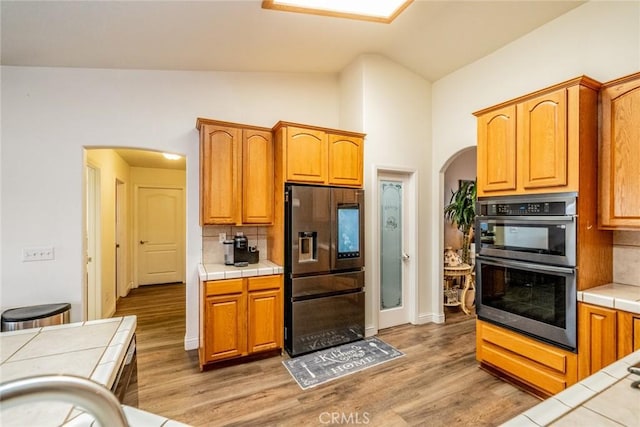 kitchen featuring tile countertops, vaulted ceiling, decorative backsplash, appliances with stainless steel finishes, and arched walkways