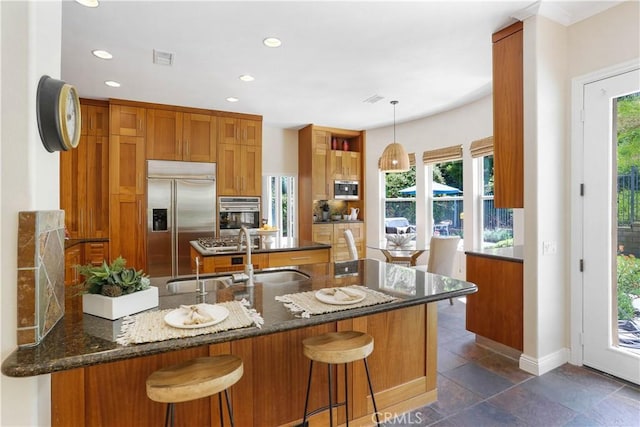 kitchen featuring stainless steel appliances, brown cabinetry, a sink, dark stone countertops, and a peninsula