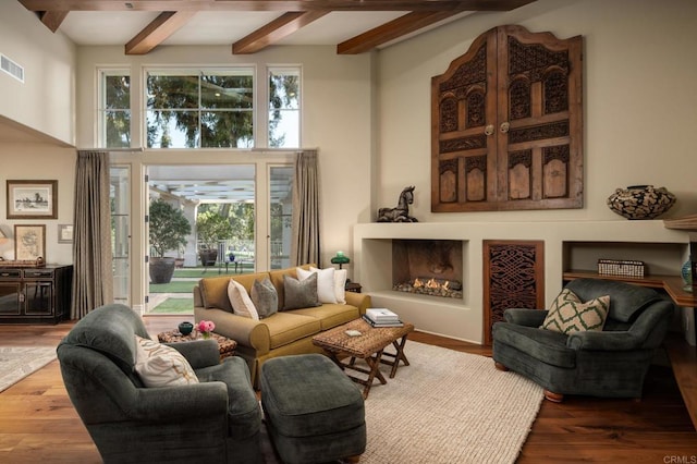 living area featuring a high ceiling, a warm lit fireplace, wood finished floors, visible vents, and beamed ceiling