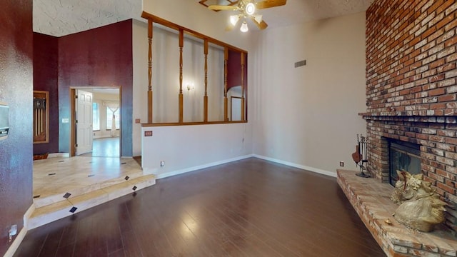 unfurnished living room with a towering ceiling, a ceiling fan, a brick fireplace, wood finished floors, and baseboards