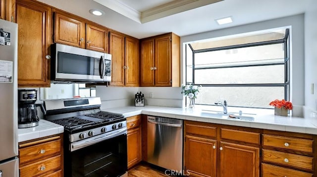 kitchen with a sink, tile counters, appliances with stainless steel finishes, a raised ceiling, and brown cabinets