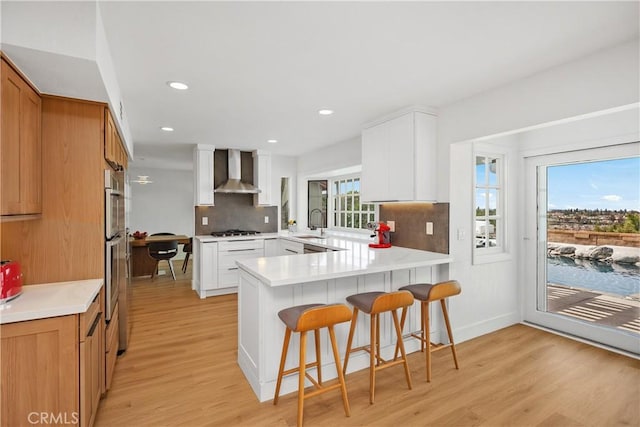 kitchen featuring a breakfast bar area, light wood-style flooring, a peninsula, a sink, and wall chimney range hood