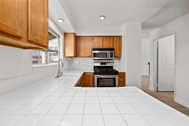 kitchen with tile counters, appliances with stainless steel finishes, brown cabinetry, and a sink