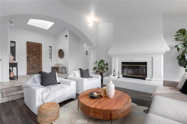 living room with vaulted ceiling with skylight, arched walkways, a tiled fireplace, and wood finished floors