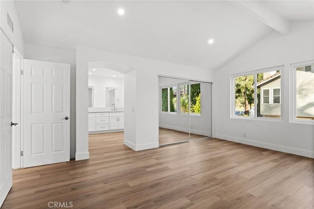 unfurnished bedroom with lofted ceiling with beams, light wood-type flooring, arched walkways, and baseboards