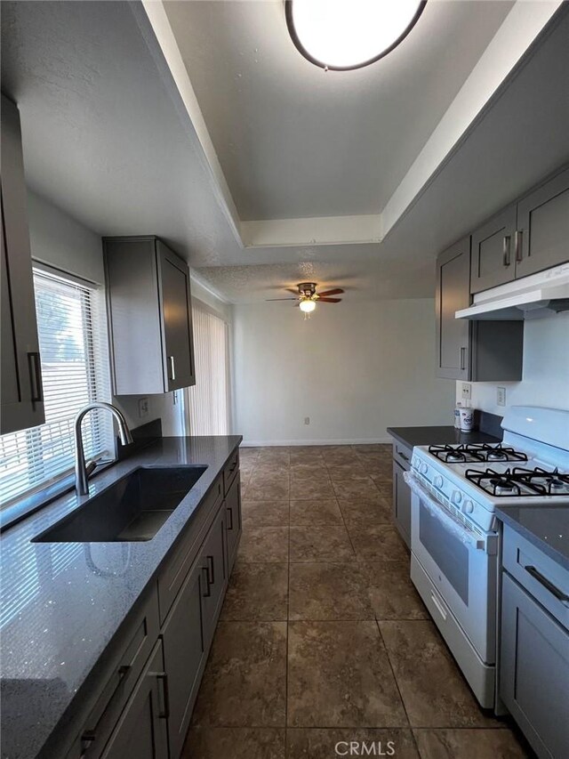 kitchen with under cabinet range hood, a tray ceiling, white gas range oven, a ceiling fan, and a sink