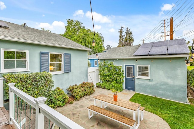 rear view of property featuring stucco siding, solar panels, a shingled roof, and a patio area