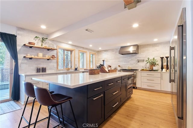 kitchen featuring light wood finished floors, white cabinets, wall chimney exhaust hood, appliances with stainless steel finishes, and light countertops