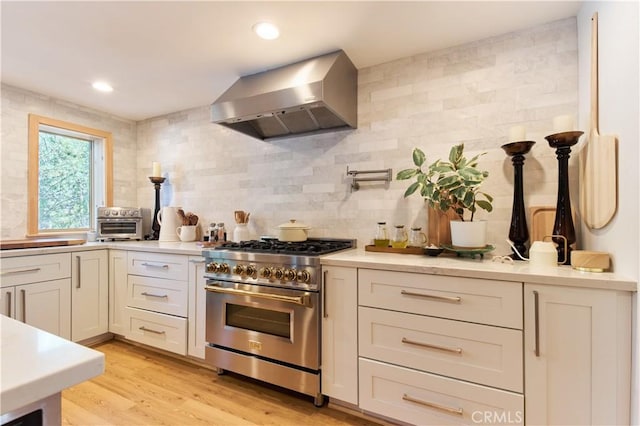 kitchen with wall chimney exhaust hood, high end range, light countertops, light wood-style floors, and backsplash