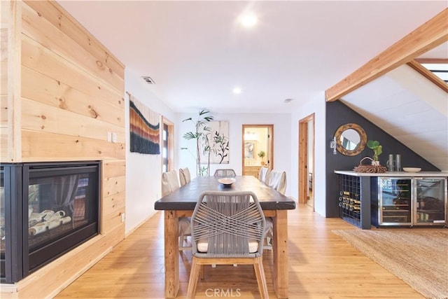 dining space featuring vaulted ceiling with beams, visible vents, a glass covered fireplace, wood finished floors, and beverage cooler