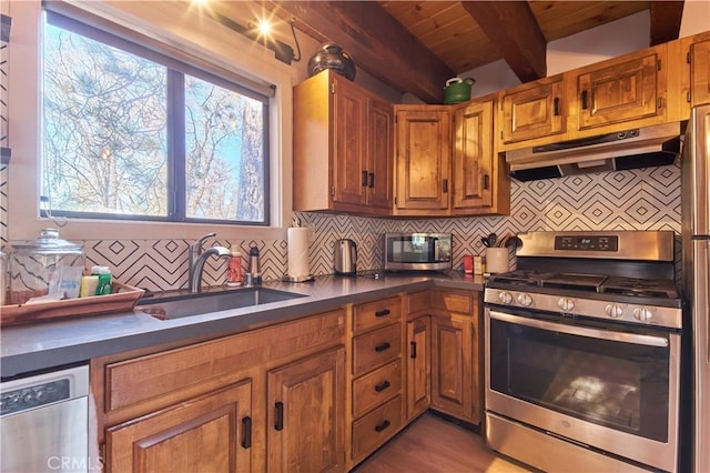 kitchen featuring under cabinet range hood, a sink, appliances with stainless steel finishes, decorative backsplash, and dark countertops