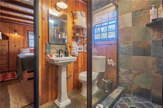 ensuite bathroom with a healthy amount of sunlight, a tile shower, toilet, and wooden walls