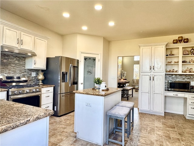 kitchen featuring appliances with stainless steel finishes, light stone counters, a center island, under cabinet range hood, and built in desk