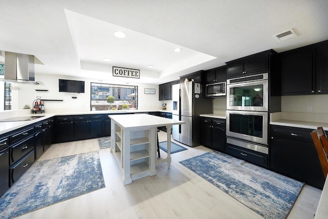 kitchen with dark cabinets, wall chimney range hood, appliances with stainless steel finishes, open shelves, and a tray ceiling