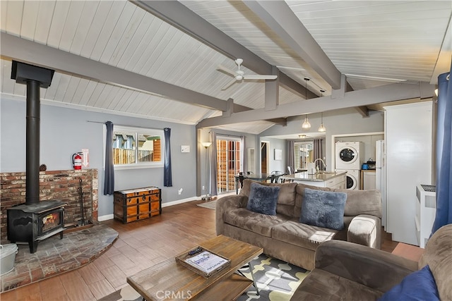 living area with baseboards, stacked washer / dryer, lofted ceiling with beams, wood finished floors, and a wood stove