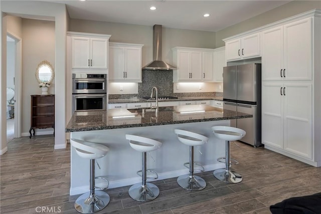 kitchen with dark wood finished floors, appliances with stainless steel finishes, a kitchen island with sink, white cabinetry, and wall chimney exhaust hood