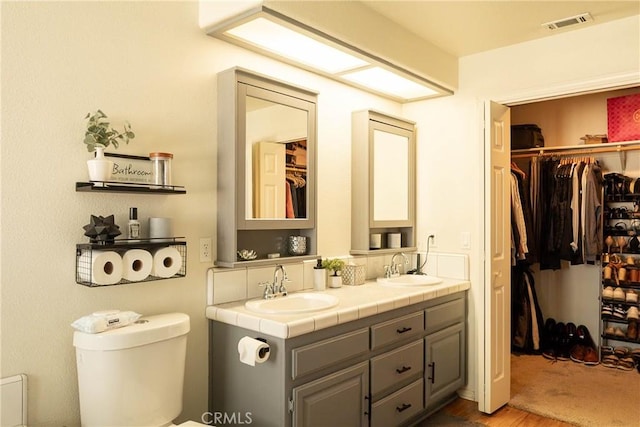 full bathroom featuring toilet, a sink, visible vents, double vanity, and a walk in closet